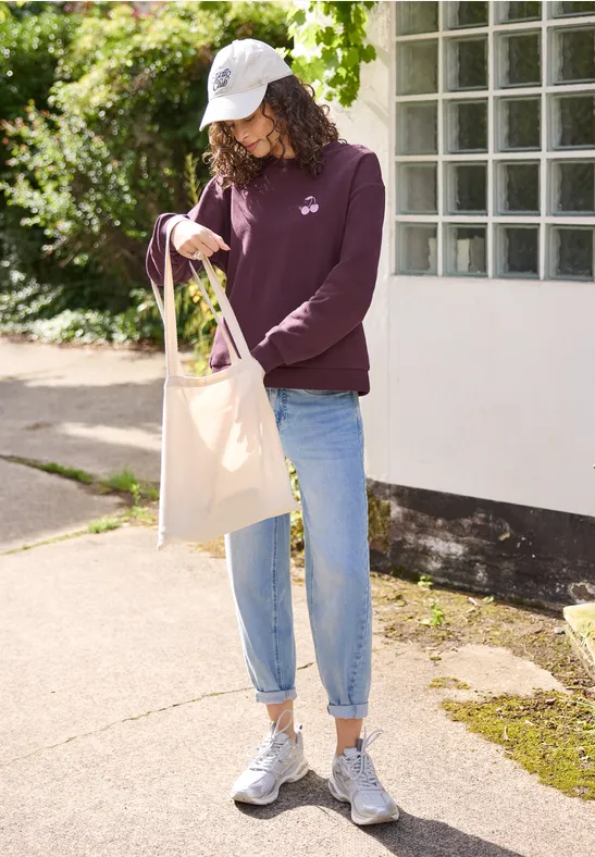 Frau mit weißer Kappe, lila Pullover und Jeans trägt eine Stofftasche und steht vor einem Haus.