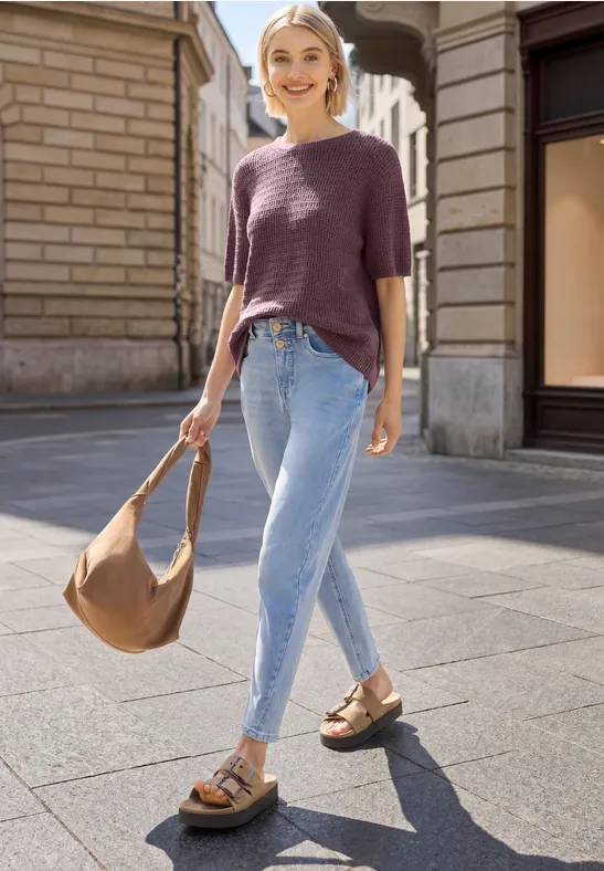 Frau in urbaner Umgebung mit lila Pullover, hellblauen Jeans, braunen Sandalen und passender Umhängetasche.