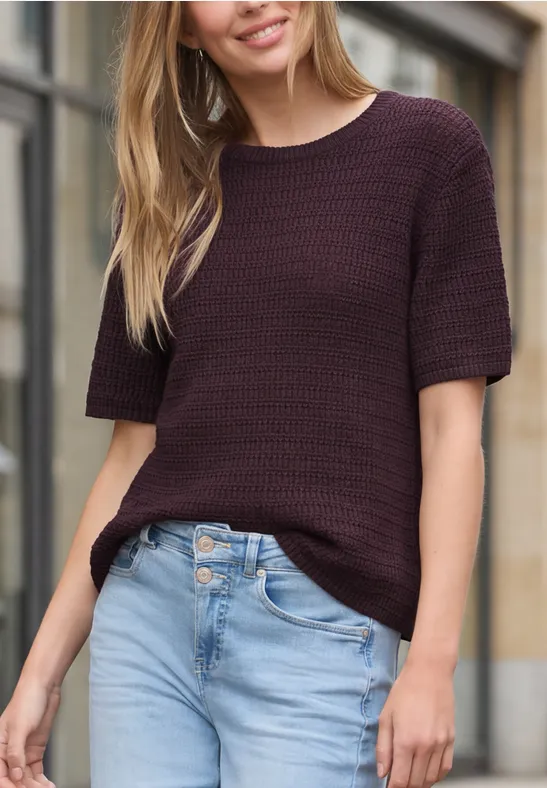 Frau in dunkelviolettem Kurzarm-Pullover und hellblauer Jeans, stehend vor einer Hausfassade.