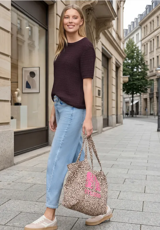 Frau in lässigem Outfit mit Strickshirt, hochgekrempelten Jeans, Slip-ons und Leoparden-Print-Tasche in Stadtgasse