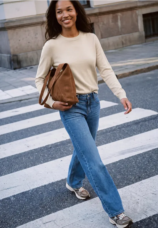 Eine lächelnde junge Frau überquert eine Straße auf einem Zebrastreifen, trägt Jeans und eine braune Tasche.