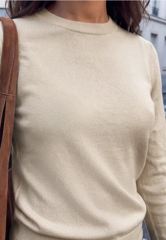 Nahaufnahme einer Person in beigem Langarm-Pullover mit brauner Ledertasche vor urbanem Hintergrund.