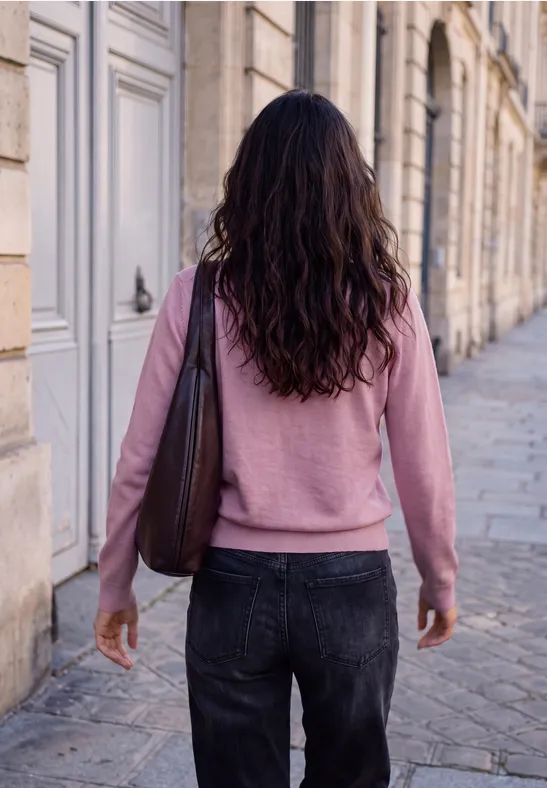 Eine Frau in rosa Pullover und dunkler Jeans mit Umhängetasche spaziert eine steinerne Straße entlang.