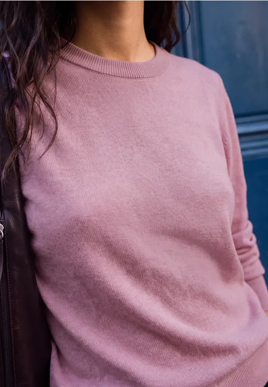 Frau mit langen dunklen Haaren trägt rosa Pullover vor dunkler Tür.