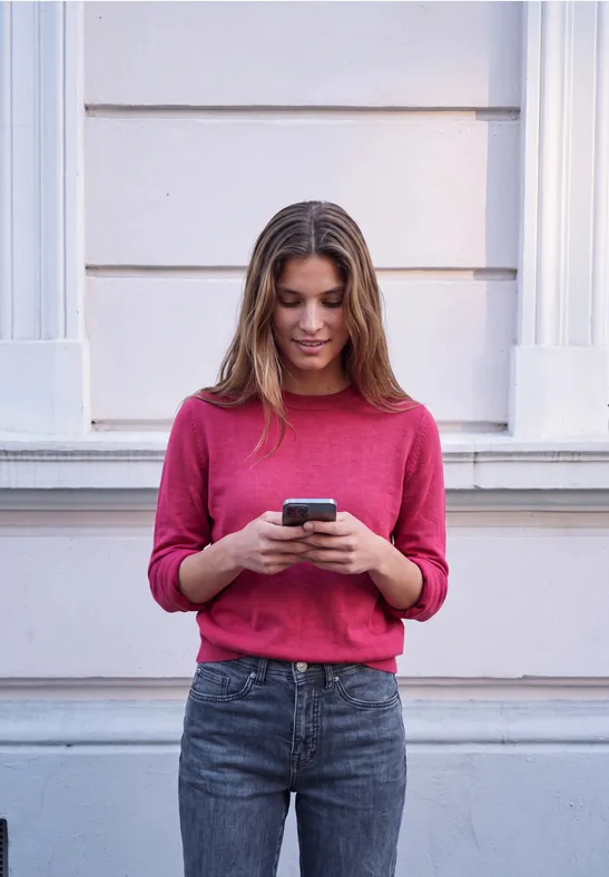 Eine junge Frau steht vor einer Hauswand und schaut auf ihr Smartphone.