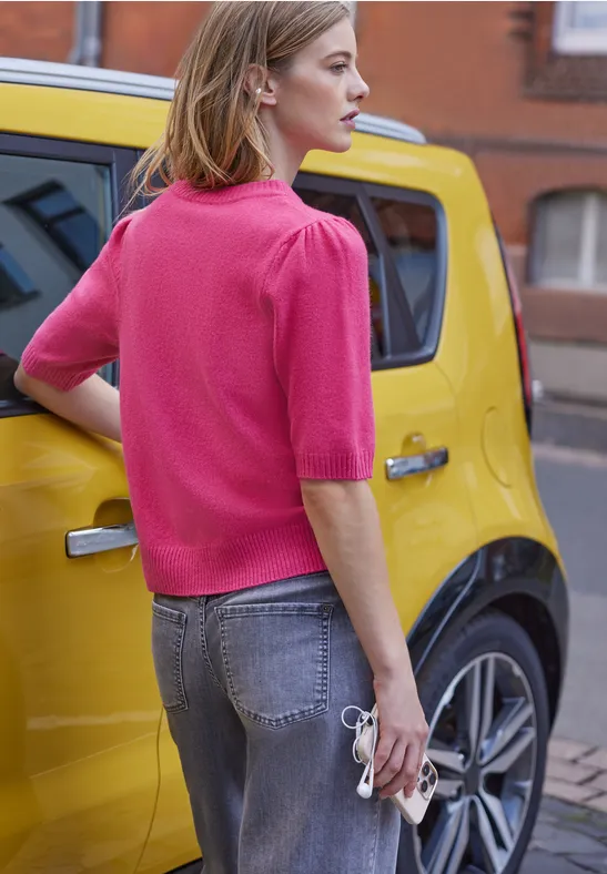 Frau mit pinkem Oberteil und Jeans hält eine Sonnenbrille neben einem gelben Auto in urbaner Umgebung.
