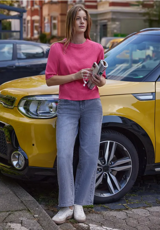 Frau in pinkem Pullover und Jeans hält eine Kamera und steht vor einem gelben Auto in der Stadt.