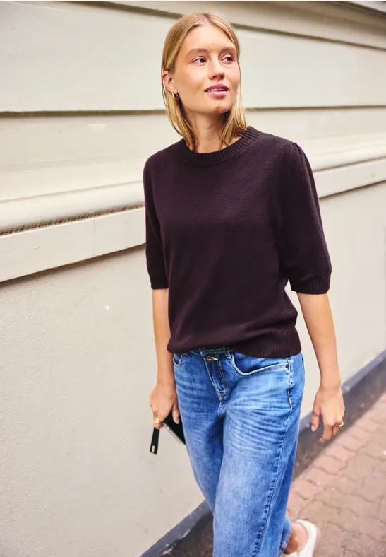 Jeune femme en t-shirt noir et jean décontracté marche devant un mur, téléphone à la main.