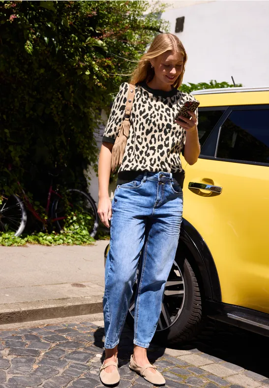 Eine Frau in Leoparden-T-Shirt und Jeans schaut neben einem gelben Auto auf ihr Smartphone.