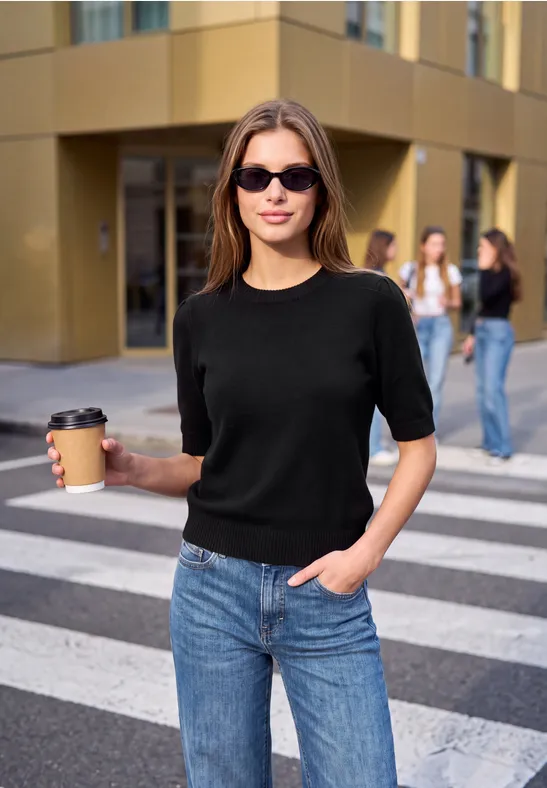 Eine Frau in Jeans und schwarzem Shirt mit Sonnenbrille hält einen Kaffee auf einem Zebrastreifen.