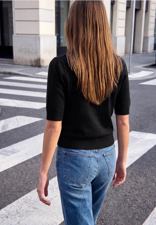 Frau in schwarzem Oberteil und Jeans überquert eine Straße an einem Zebrastreifen in einer Stadt.