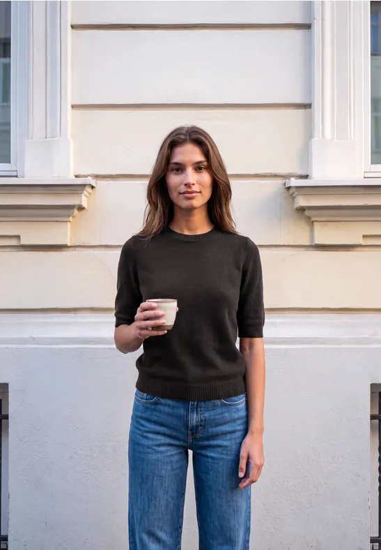 Młoda kobieta stoi przy elewacji, trzyma kubek kawy, ma na sobie czarny T-shirt i dżinsy.