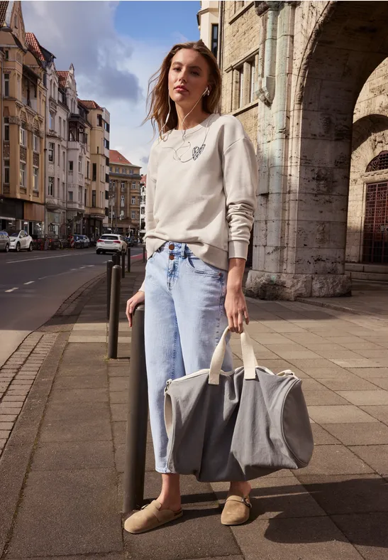 Osoba z niewyraźną twarzą w dżinsach i swetrze stoi przed starymi kamienicami, trzymając dużą torbę.