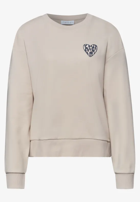 Jasnobeżowa bluza z okrągłym dekoltem, długimi rękawami i małą naszywką w kształcie serca na piersi.