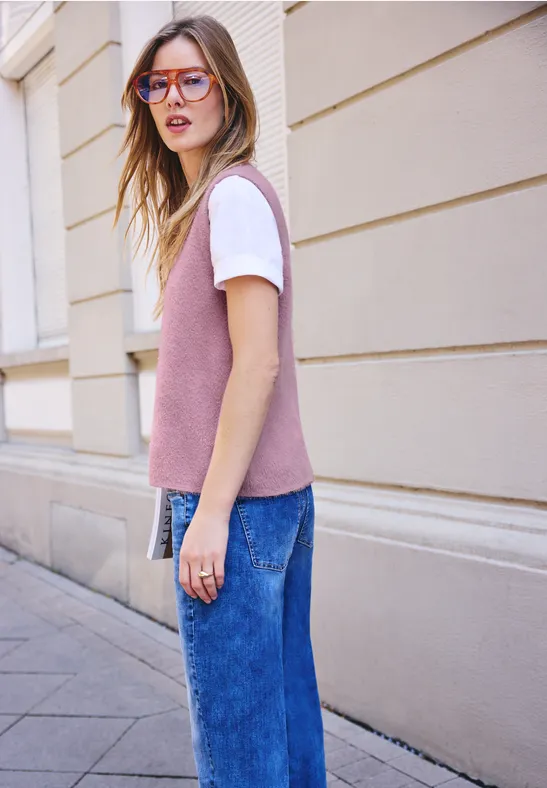 Frau mit rosa Pullunder, weißem T-Shirt, blauen Jeans und Brille steht auf einem Gehweg vor einem Gebäude.