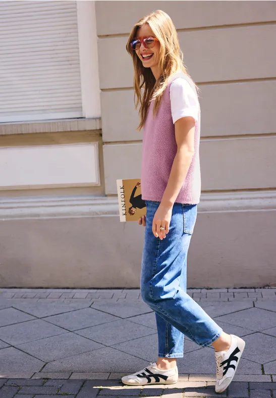 Eine lächelnde junge Frau in Jeans, rosa Pullunder und Sonnenbrille geht auf dem Bürgersteig entlang.