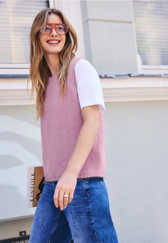 Eine Frau mit rosa Pullunder, weißem T-Shirt und Sonnenbrille geht lächelnd spazieren.