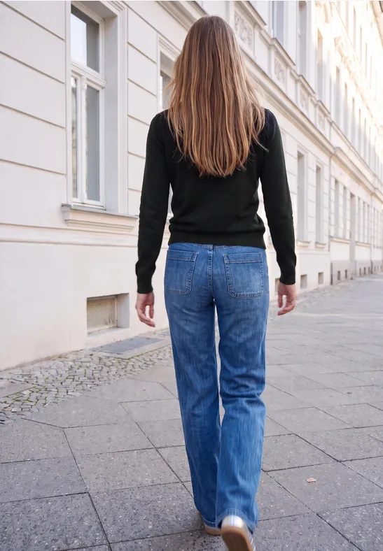 Frau mit langen Haaren in Jeans und schwarzem Pullover läuft auf einem städtischen Bürgersteig.