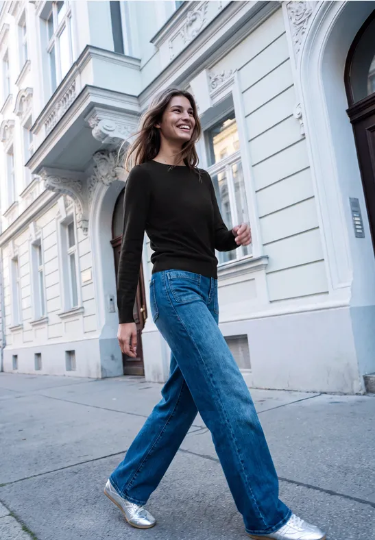Junge Frau in schwarzem Pullover und blauen Jeans spaziert selbstbewusst eine Straßenfassade entlang.