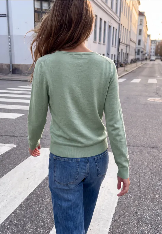 Rückansicht einer Person in grünem Pullover und blauer Jeans, die über eine ruhige Straße geht.