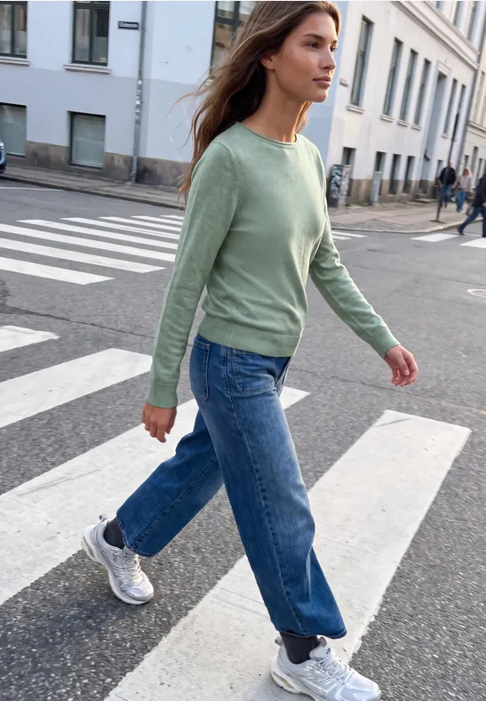 Eine junge Frau in grüner Strickjacke, Jeans und Sneakers überquert einen Zebrastreifen in der Stadt.