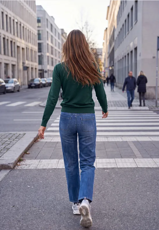 Eine Person mit grünem Pullover und Jeans überquert einen Zebrastreifen in der Stadt.