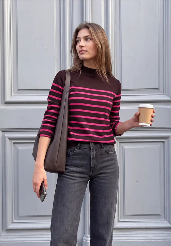 Une jeune femme devant une porte, tenant un café et un smartphone, portant un pull rayé et un jean.