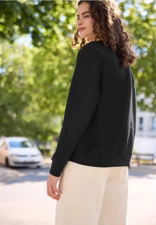 Junge Frau mit lockigem Haar trägt schwarzen Pullover und beigefarbene Hose, steht im Freien.