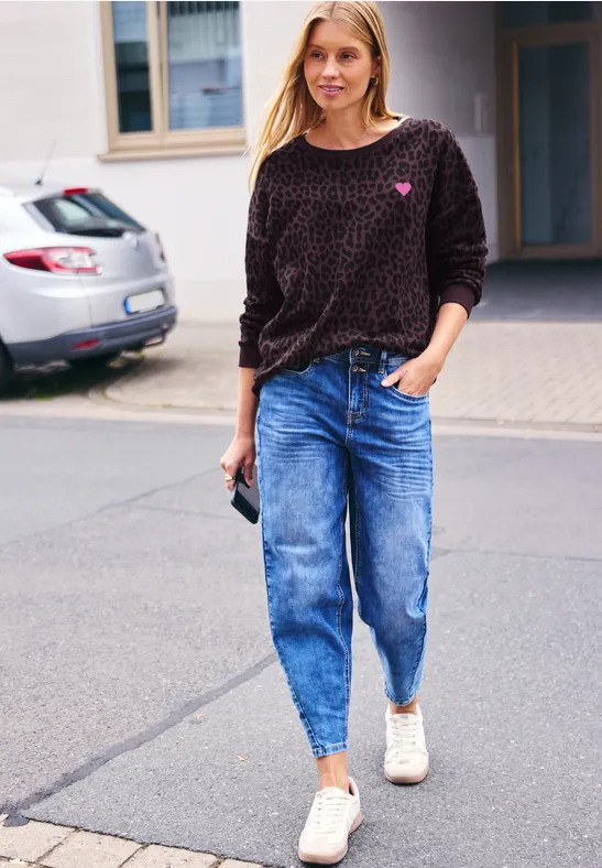 Eine Frau trägt Jeans, gemusterten Pullover und weiße Sneakers, läuft auf der Straße und hält ein Handy.