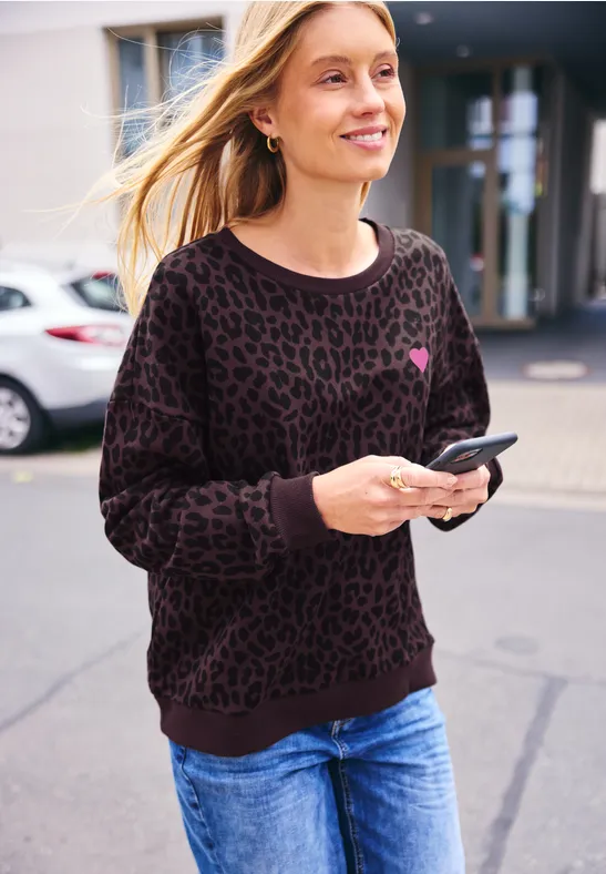 Kobieta w dżinsach i leopardim swetrze idzie ulicą, uśmiecha się i trzyma smartfon.