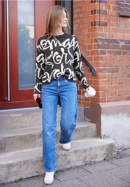 Frau verlässt ein Backsteinhaus mit Kaffebecher und Schlüssel, trägt schwarzen Pullover und blaue Jeans.
