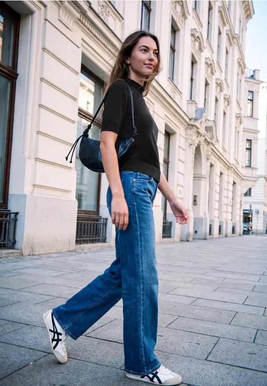 Frau in schwarzem Shirt und blauer Jeans spaziert an historischen Stadthäusern vorbei.