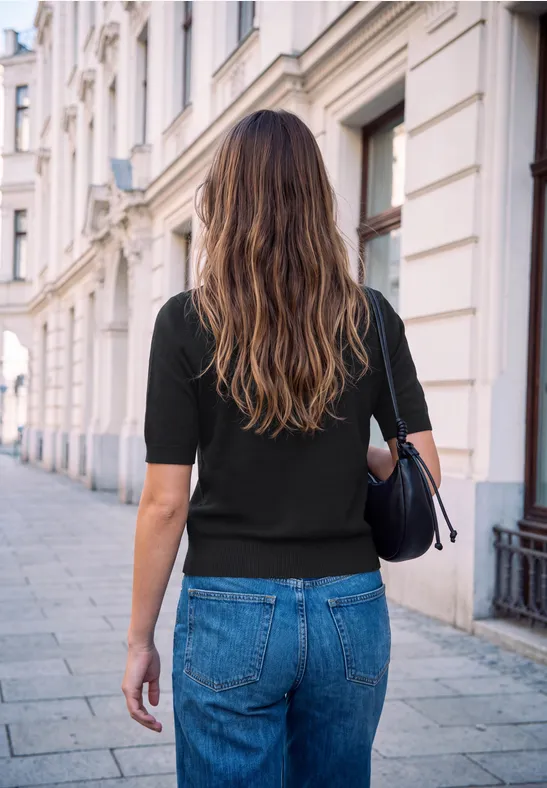 Eine Frau mit langem Haar, schwarzem Shirt und Jeans spaziert auf einer gepflasterten Straße vor historischen Häusern.