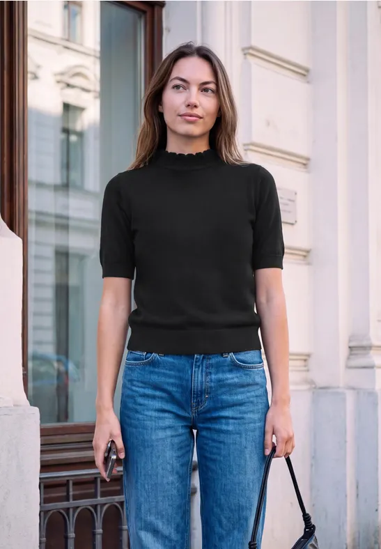 Eine Frau in Jeans und schwarzem Oberteil steht vor einem Gebäude, hält ein Handy und eine Tasche.
