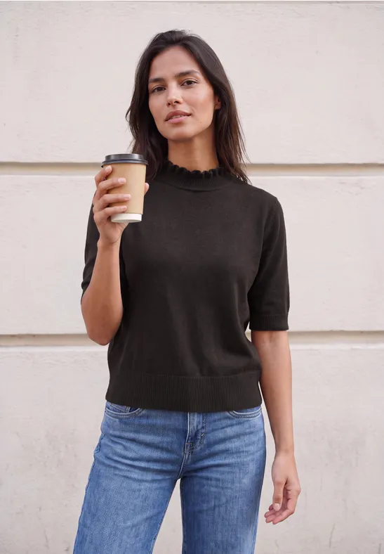 Une femme devant un mur, en pull noir et jean, tenant un gobelet de café à emporter.