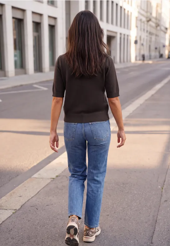 Femme en jean et haut noir se promène dans une rue de la ville ensoleillée.