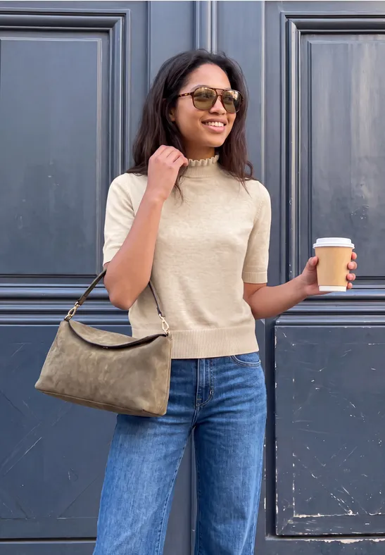 Frau mit Sonnenbrille, hellem Strickoberteil und Jeans trägt Handtasche und hält Kaffee.