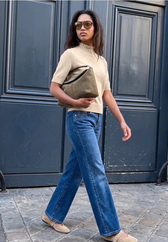 Frau mit Brille, beige Pullover, blauen Jeans und Handtasche schreitet vor dunkler Tür entlang