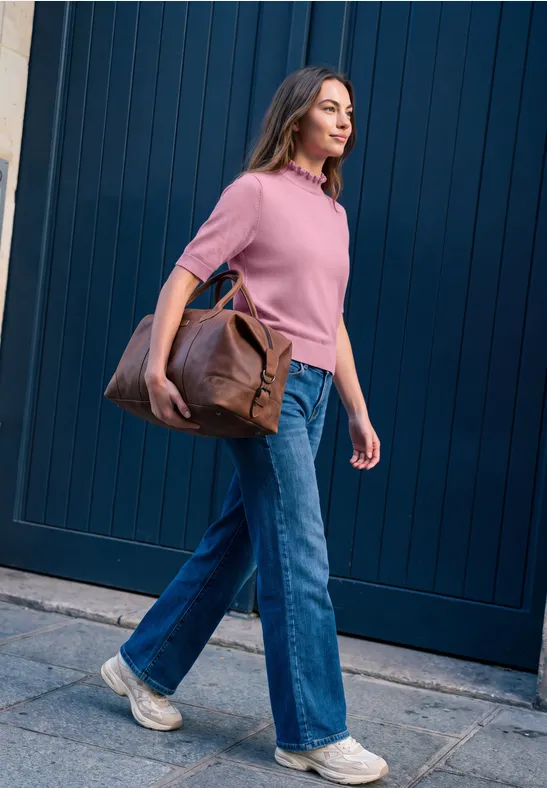 Frau in rosa Pulli, blauen Jeans und Sneakers geht an einer dunkelblauen Wand entlang und trägt eine braune Tasche.