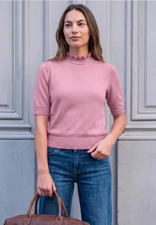 Frau im rosa Kurzarm-Pullover und Jeans hält Handtasche vor hellgrauem Hintergrund.