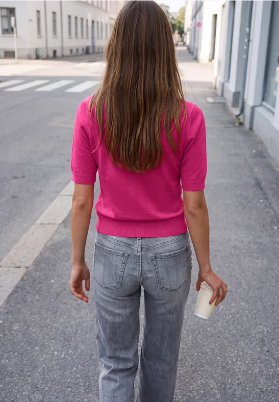 Frau von hinten mit langem Haar, pinkem Oberteil und grauen Jeans, die einen Pappbecher auf der Straße hält.