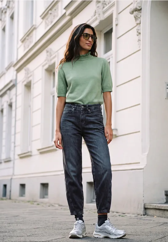 Frau in grünem Pullover und Jeans steht auf dem Gehweg vor einem Gebäude.