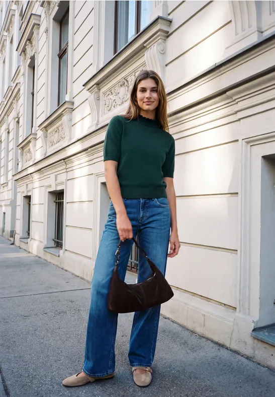 Eine lächelnde Frau in grünem Oberteil, blauen Jeans und beigen Flats steht mit Handtasche vor Hausfassade.