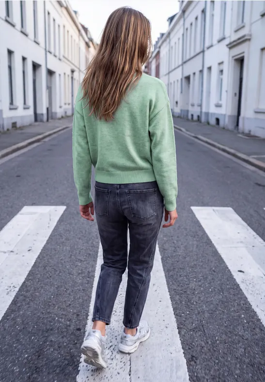 Een vrouw met een groen truitje, donkere spijkerbroek en witte sneakers steekt een rustige woonstraat over.