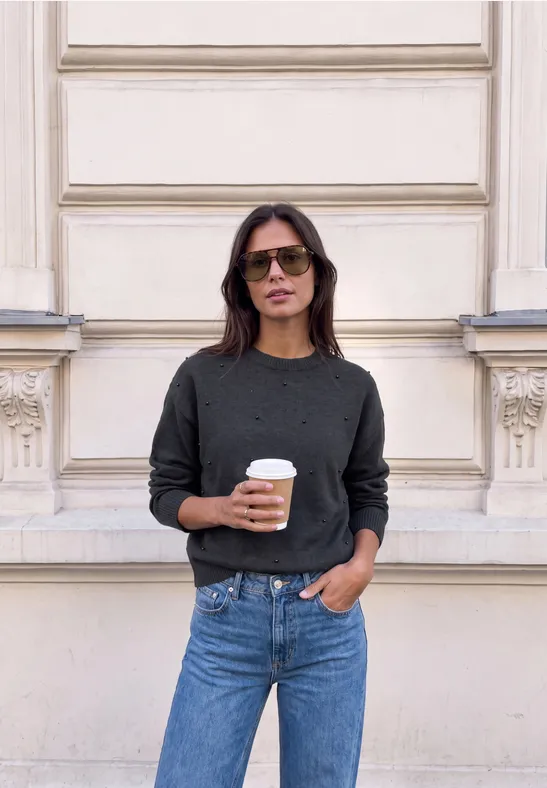 Frau mit Sonnenbrille und Kaffee vor heller Hausfassade.