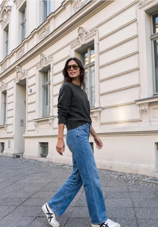 Frau in schwarzem Pullover, Jeans und weißen Sneakern spaziert vor historischem Gebäude.