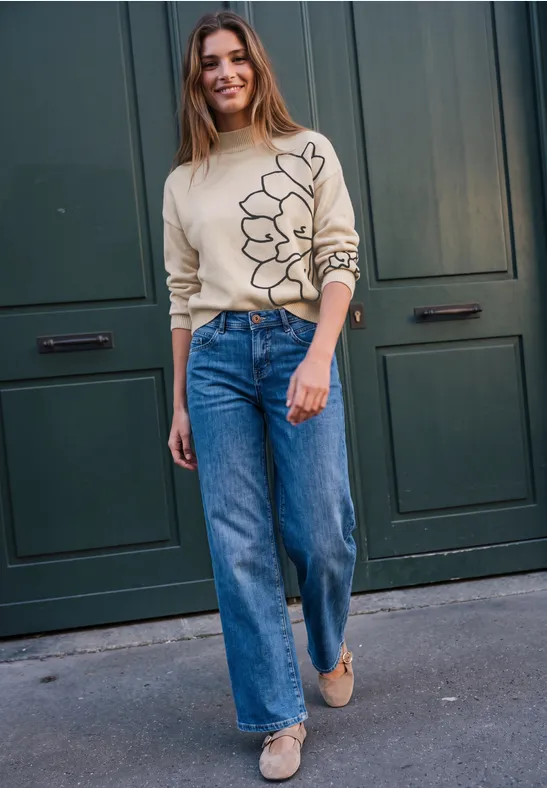 Junge Frau in beigem Pullover mit Blumenmotiv und weiten Jeans steht lächelnd vor einer grünen Tür.