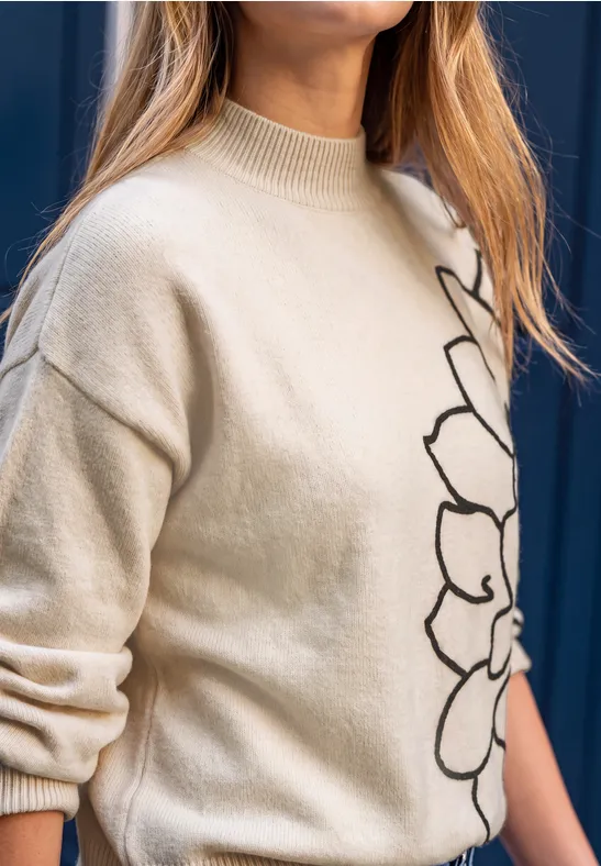 Frau im beigen Strickpullover mit schwarzem Blumenmuster vor dunkelblauem Hintergrund.