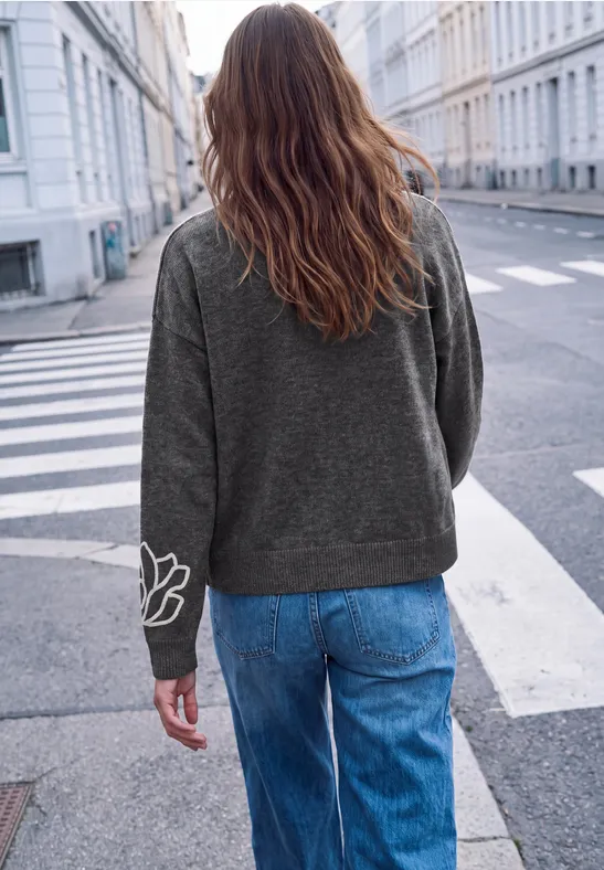 Person mit langen Haaren in grauem Pullover und blauen Jeans steht auf einem Zebrastreifen in einer Stadt