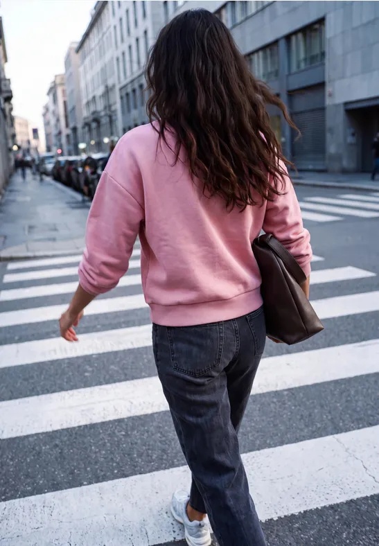 Eine Frau in rosa Pullover und dunklen Jeans überquert eine Straße an einem Zebrastreifen in der Stadt.
